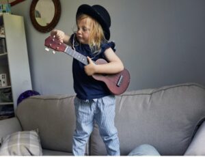 Little girl wearing a hat standing on a couch with a ukulele