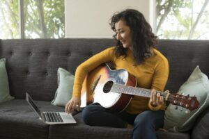 Young woman in a yellow shirt sitting down watching a video of how to play the guitar