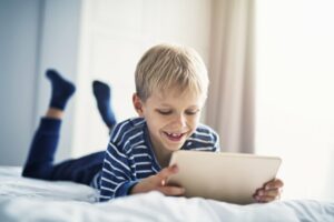 Smiling little boy laying on the bed playing on his tablet