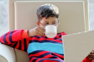 Young boy wearing glasses sipping from a mug reading newspaper